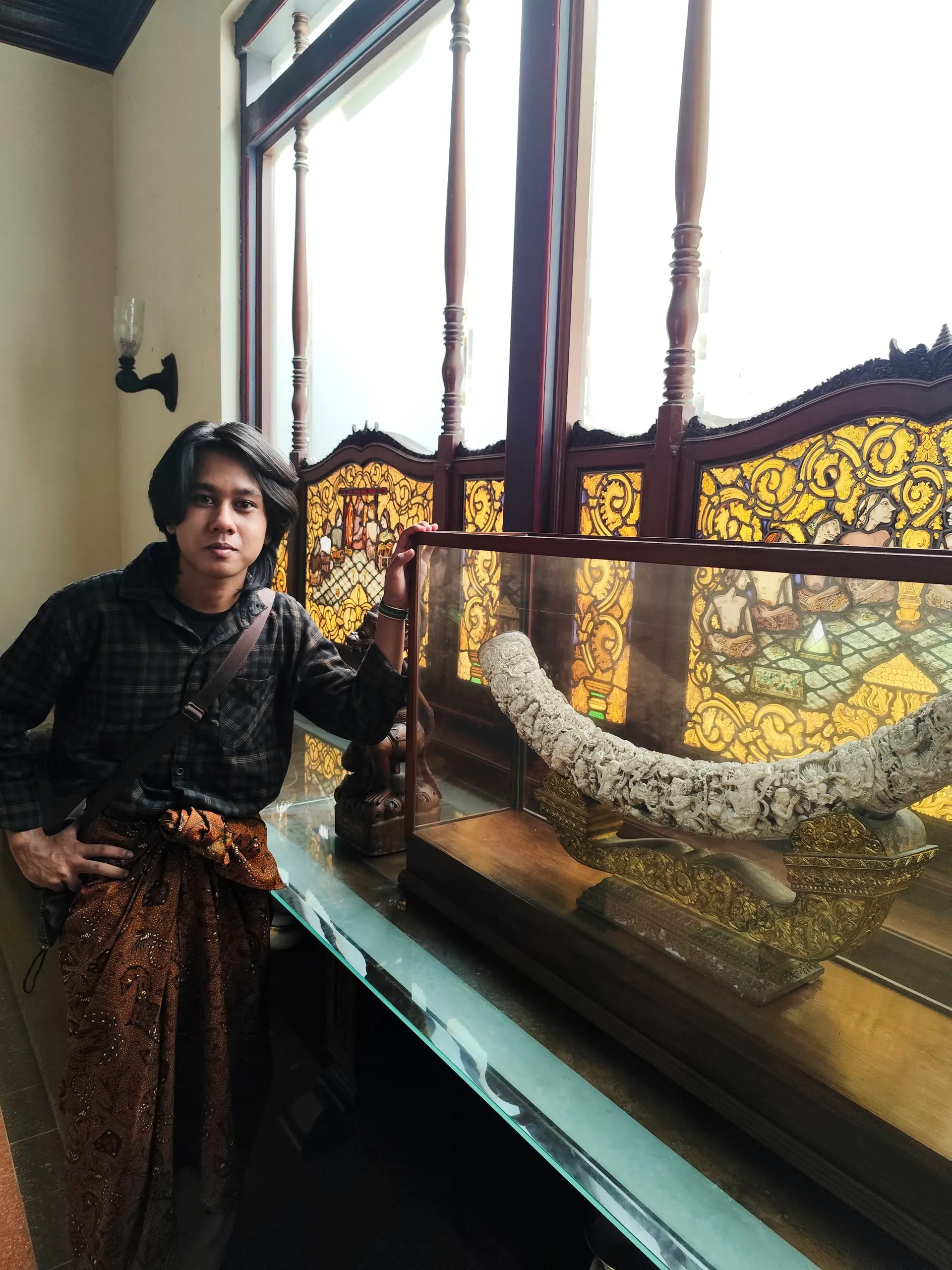 Portrait of Azril in a batik sarong next to a carved display case in Solo